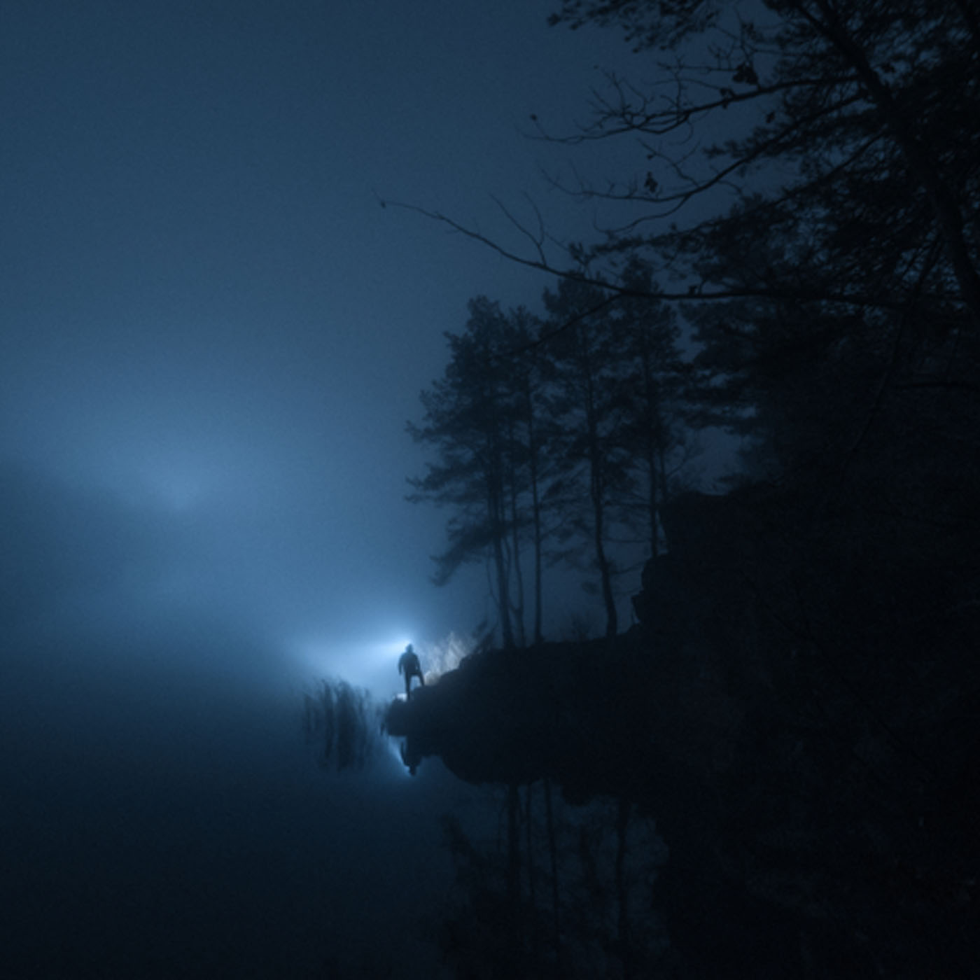 Silhouette of a person with flashlight by foggy lake at night