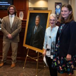 Jack Rizzo '25, Betsy Calvo Anderson, and Lily Frautschi J.D. '26 pose with the portrait of the late Alvin P. Anderson at TowneBank.