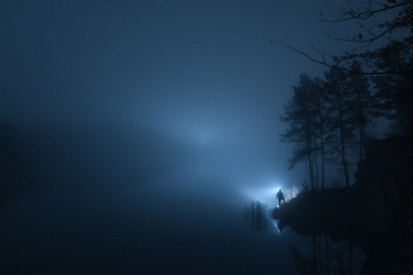 Silhouette of a person with flashlight by foggy lake at night